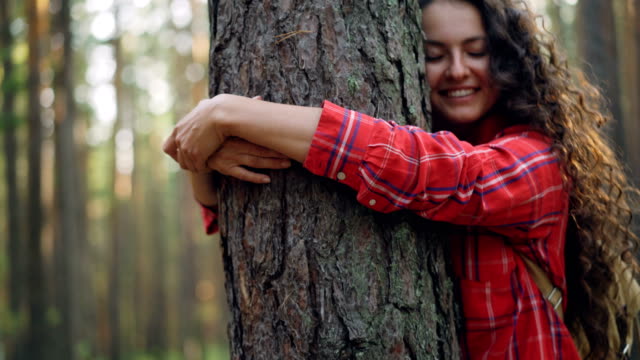 Hermosa-mujer-joven-con-pelo-rizado-con-camisa-brillante-es-abrazar-árboles-disfrutando-de-la-naturaleza-y-sonriendo-con-los-ojos-cerrados.-Concepto-personas,-recreación-y-felicidad.