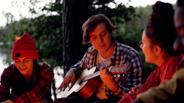 Handsome-young-man-tourist-is-playing-the-guitar-and-smiling-while-his-friends-are-singing-and-having-fun-resting-around-fire-near-lake-or-river.-Green-trees-are-visible.