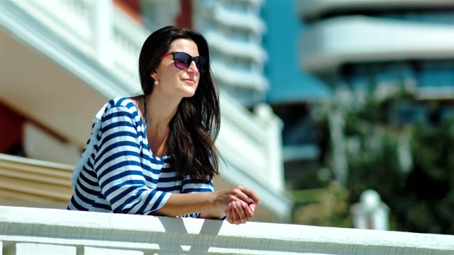 Glücklich-lächelnd-weibliche-Touristen-mit-Sonnenbrille-und-gestreiften-Kleid-legte-Hände-auf-weißen-Geländer