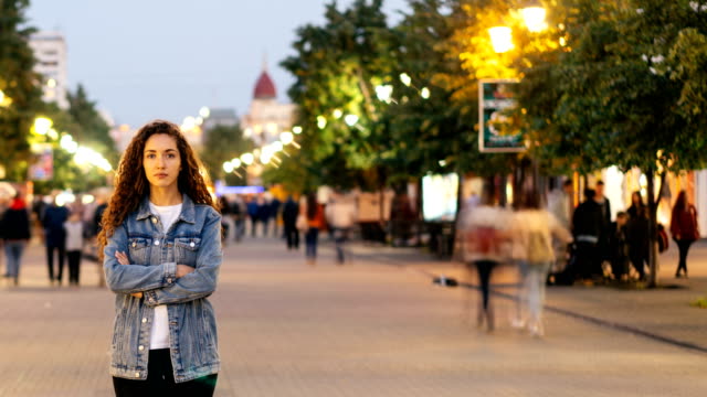 Time-lapse-of-unhappy-girl-standing-alone-on-beautiful-pedestrian-street-late-in-the-evening-and-looking-at-camera-when-people-are-whizzing-around.-Society-and-loneliness-concept.