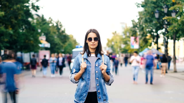 Time-lapse-portrait-of-attractive-brunette-in-sunglasses-standing-in-the-street-by-herself-and-looking-at-camera-when-men-and-women-are-passing-by-on-autumn-day.