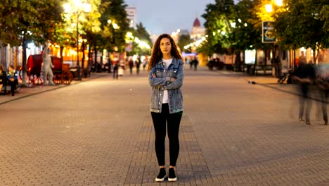 Time-lapse-of-attractive-woman-with-long-curly-hair-standing-in-the-street-with-crossed-arms-and-looking-at-camera-on-autumn-evening-when-people-are-moving-around.