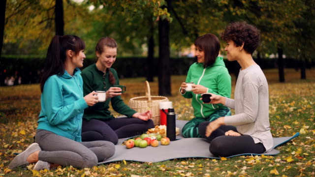 Happy-girls-are-having-picnic-in-park-sitting-on-yoga-mats-and-eating-after-outdoor-practice-in-autumn,-girls-are-talking-and-laughing.-Communication-and-food-concept.