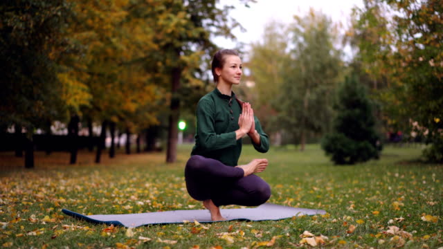 Beautiful-girl-yogini-is-exercising-in-park-on-mat-practising-balancing-positions-on-one-leg-and-relaxing.-Healthy-lifestyle,-free-time-activity-and-people-concept.