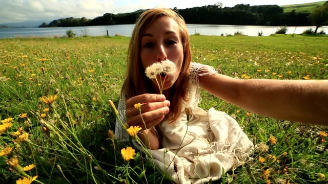 Female-takes-selfie-portrait-while-blowing-flower's-seeds-in-meadow