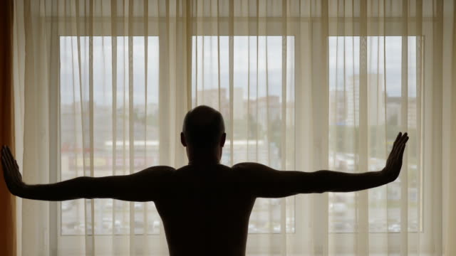 Man-infront-of-curtains-in-the-morning-raises-his-hands-and-stretch-oneself.-Adult-man-and-big-window-with-curtains.