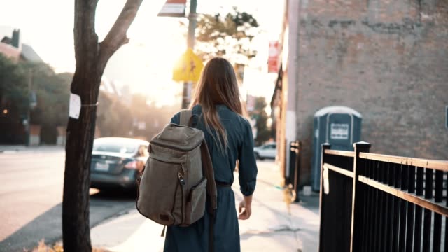 Back-view-of-young-stylish-woman-with-backpack-walking-alone-on-sunset-in-summer,-spending-time-in-downtown