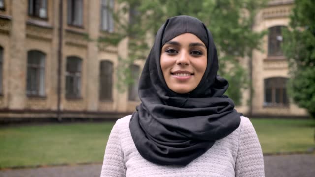 Junges-schönes-muslimische-Mädchen-Hijab-ist-stehend-und-lächelnd-in-tagsüber-bei-schlechtem-Wetter,-gerade-in-die-Kamera,-aufbauend-auf-Hintergrund,-Religiuos-Konzept
