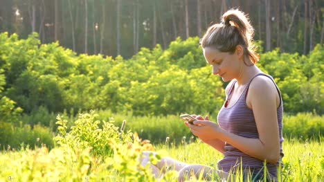 Glückliches-Mädchen-sitzen-auf-grünem-Rasen-und-nutzt-Smartphone-auf-szenischen-Bereich-bei-Sonnenuntergang-Hintergrund