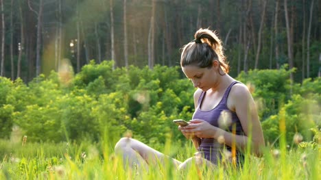 Glückliche-junge-Frau-sitzt-auf-dem-grünen-Rasen-und-nutzt-Smartphone-auf-malerischen-Feld-bei-Sonnenuntergang-Hintergrund
