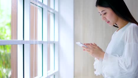 schöne-asiatische-Frau-genießen-Glück-Hand-Verwendung-Smartphone-in-der-Nähe-von-Fenster