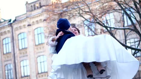 winter-wedding.-newlywed-couple-in-wedding-dresses.-groom-holds-bride-in-his-arms,-spinning.-they-are-happy,-smiling-at-each-other.-background-of-ancient-architecture,-snow-covered-park
