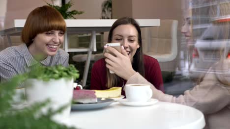 Drei-junge-kaukasische-Mädchen-sitzen-im-Café,-lachen,-Lächeln,-Freunde,-Firma,-Klatsch,-Dialog,-Diskussion.-Freundinnen-in-der-Café-Konzept.-Sehen-Sie-ein-lustiges-Video,-ein-Foto-mit-einem-Smartphone-60-fps