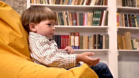 Kleiner-Junge-sitzt-auf-Sessel-und-fangen-und-wirft-Ball,-Bücherregale-Hintergrund