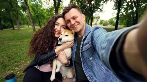 Point-of-view-shot-of-two-happy-people-and-puppy-taking-selfie-in-the-park,-kissing-and-loving-expressing-love-and-care.-Beautiful-green-lawn-and-trees-are-in-background.