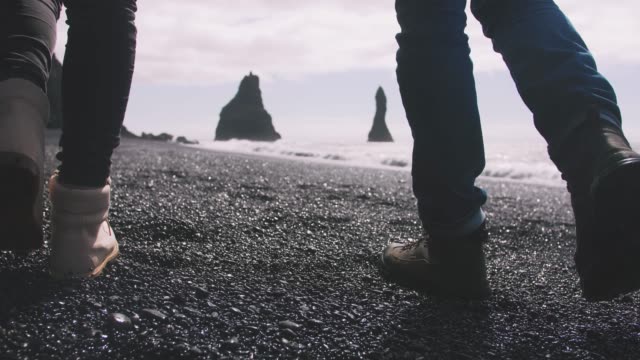Cerrar-tiro-de-joven-pareja-cogidos-de-la-mano-caminando-por-la-playa-de-arena-negra-en-Islandia,-cámara-lenta