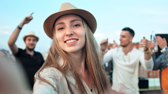 Beautiful-European-woman-in-hat-taking-selfie-using-camera-or-smartphone-at-summer-rooftop-party