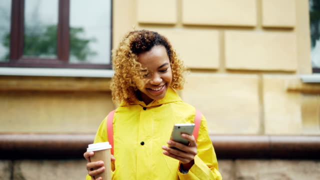 Ziemlich-afroamerikanische-Mädchen-ist-Smartphone-Bildschirm-betrachten-und-berühren-mit-Lächeln-stehen-im-freien-helle-Kleidung-trägt-und-hält-Kaffee.-Mensch-und-Technik-Konzept.