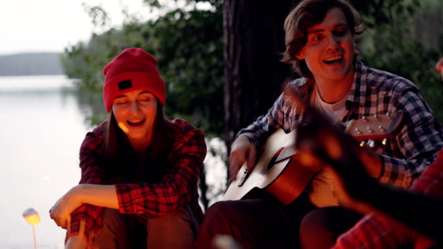 Multiracial-group-of-tourists-is-having-fun-in-forest-playing-the-guitar-and-singing-songs-around-campfire-in-wood-near-lake.-People-are-wearing-casual-clothing.