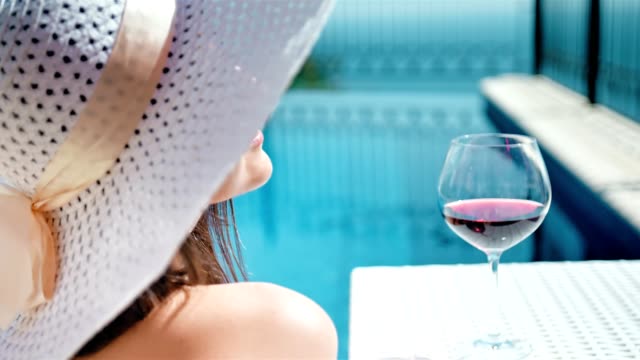 Portrait-smiling-pretty-woman-in-floppy-hat-and-sunglasses-enjoying-sunbathing-with-goblet-of-wine