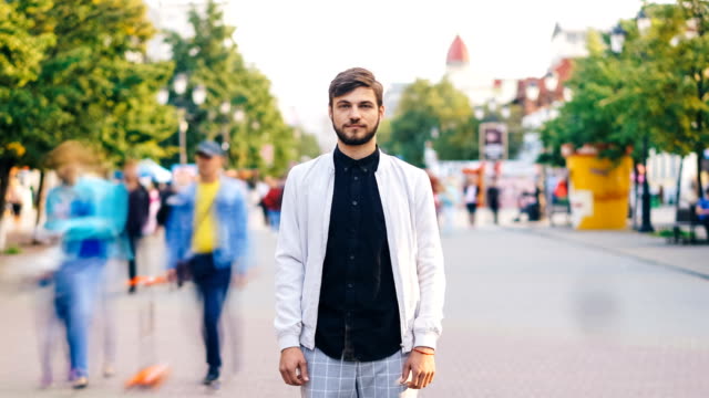 Time-lapse-portrait-of-serious-young-man-looking-at-camera-standing-in-center-of-busy-pedestrian-street-in-summer-wearing-stylish-clothing-while-people-are-passing-by.