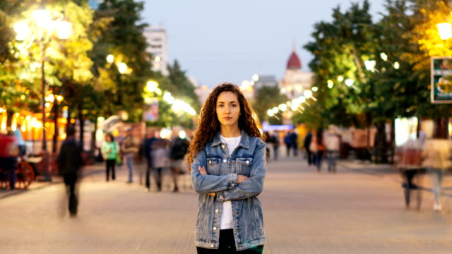 Time-lapse-of-good-looking-woman-towny-in-denim-clothing-standing-alone-in-the-street-among-crowds-of-people-and-looking-at-camera.-Street-lights-are-visible.