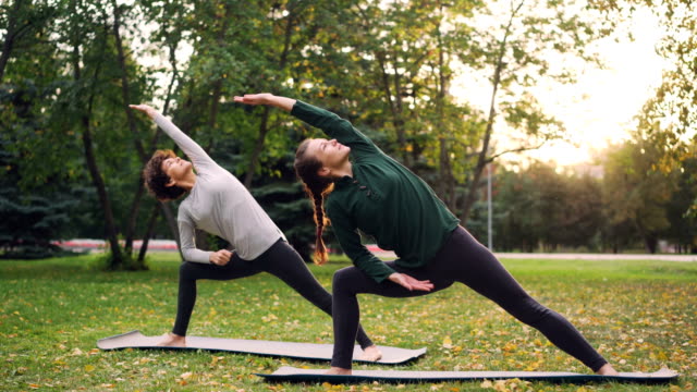 Two-pretty-women-are-doing-yoga-outdoors-in-park-on-mats-practising-asanas-and-breathing-fresh-air.-Individual-practice,-professional-instructor-and-nature-concept.