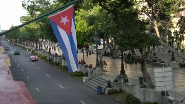 high-angle-aerial-establishing-shot-of-classic-convertible-car-on-street-with-cuban-flag-in-Havana,-Cuba