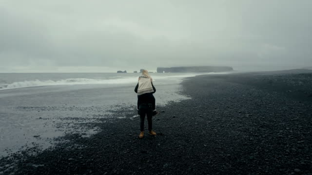 Aerial-view-of-the-young-couple-walking-on-the-black-volcanic-beach-in-Iceland.-Woman-in-lopapeysa-jumps-on-the-man-arms