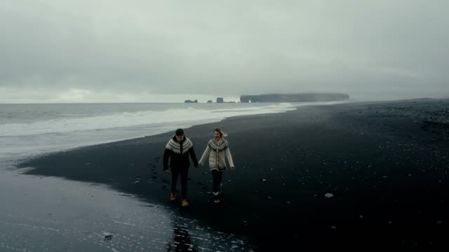 Aerial-view-of-young-couple-walking-on-the-shore-of-the-sea-in-Iceland.-Man-and-woman-look-to-the-wave-at-black-beach