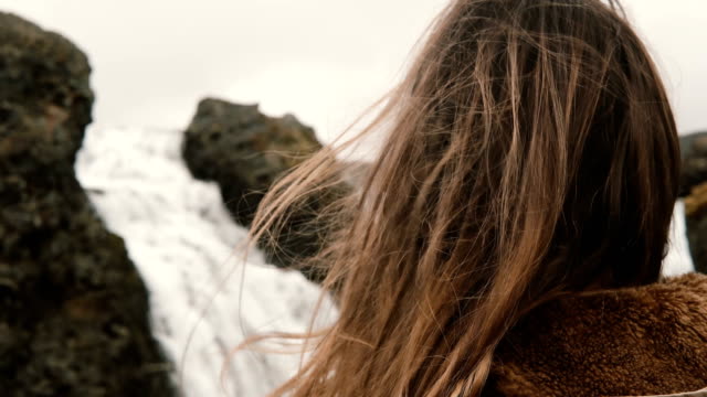 Portrait-of-young-beautiful-sad-woman-standing-near-the-waterfall-in-Iceland-and-looking-around.-Hair-waving-on-wind