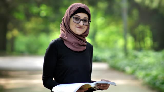 Retrato-de-una-joven-con-gafas-en-un-hiyab-leyendo-un-libro-en-la-naturaleza,-riendo,-en-un-parque-en-el-fondo.-50-fps