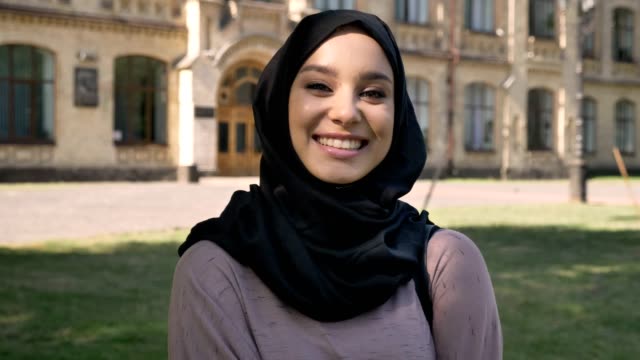 Young-beautiful-muslim-girl-in-hijab-is-standing-and-smiling-in-daytime-in-summer,-watching-at-camera,-building-on-background,-religiuos-concept