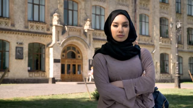 Young-serious-muslim-girl-is-standing-with-arms-crossed-in-daytime-in-summer,-watching-at-camera,-building-on-background,-religious-concept