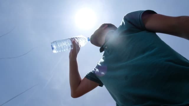 Young-man-drinking-bottle-of-water-standing-outdoor-in-hot-summer