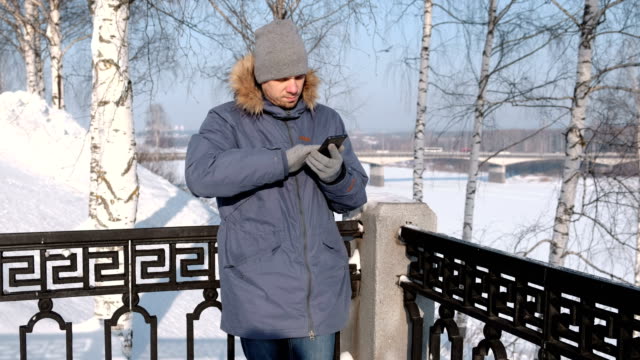 Mann-in-blauer-Jacke-mit-Fell-Kapuze-wischt-den-Handy-Bildschirm-mit-seiner-Hand-in-Handschuhe-im-Winter-Park.