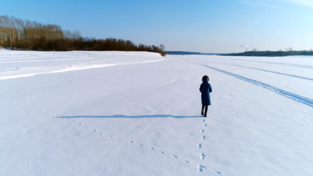 Luftaufnahmen-von-laufenden-Frau-in-Winterlandschaft-eines-gefrorenen-Flusses-mit-einem-Wald-und-einem-Blick-auf-die-Stadt-mit-Fabriken.-Hinten-Sie-Ansicht-von.