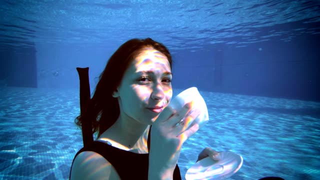A-charming-young-girl-is-sitting-underwater-at-the-bottom-of-the-pool-in-a-beautiful-dress,-holding-a-white-Cup-and-saucer-in-her-hands,-looking-at-the-camera-and-smiling.-Vintage-processing.