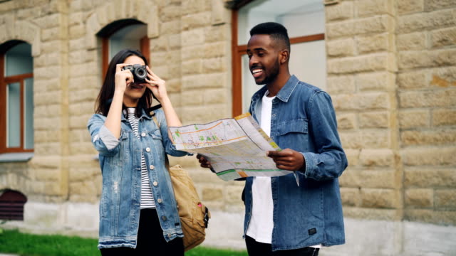 Happy-African-American-guy-and-Caucasian-girl-travelers-are-looking-at-map-and-taking-photos-standing-outdoors-then-leaving.-Multiracial-friendship-and-tourism-concept.