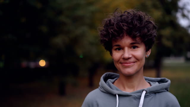 Portrait-of-good-looking-girl-with-short-curly-hair-looking-at-camera-and-smiling-then-laughing-standing-outdoors-in-autumn-in-city-park.-People-and-nature-concept.