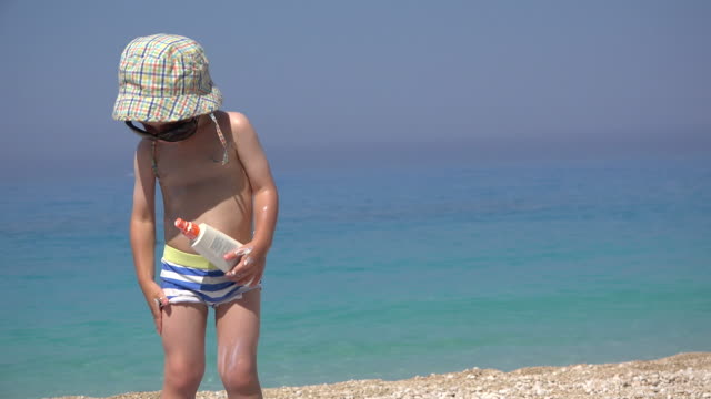Lustiges-kleines-Kind-mit-Sonnenbrille-und-Hut,-Sonnencreme-auf-seinem-Körper-anwenden