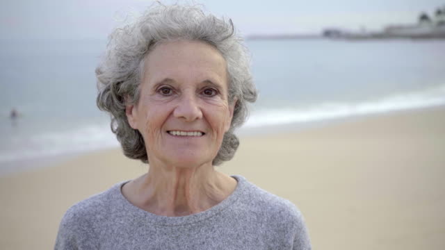Happy-beautiful-elderly-woman-with-toothy-smile-posing-against-blurred-seascape-background.