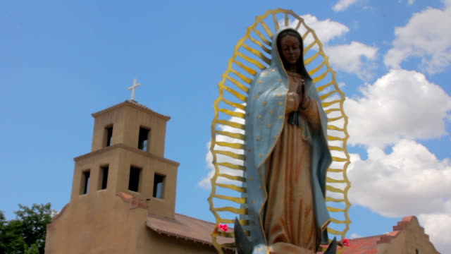 Panning-shot-de-una-estatua-de-nuestra-Señora-de-Guadalupe,-frente-a-una-iglesia-de-Adobe