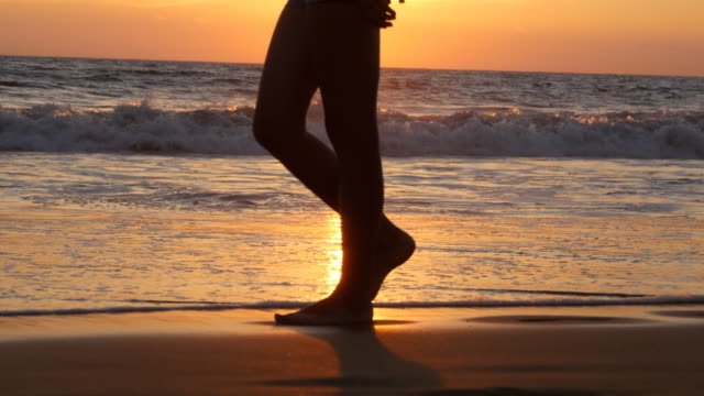 Legs-of-young-woman-going-along-ocean-beach-during-sunrise.-Female-feet-walking-barefoot-on-sea-shore-at-sunset.-Girl-stepping-in-shallow-water-at-shoreline.-Summer-vacation-concept.-Close-up
