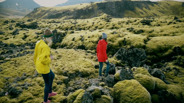Vista-aérea-de-las-dos-mujeres-caminando-en-el-campo-de-lava-en-Islandia.-Los-turistas-toca-las-protuberancias-de-musgo