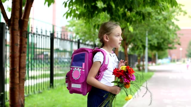 Schoolgirl-with-backpack,-flowers-bouquet-say-goodbye-and-go-away.-Girl-standing-near-school-with-bouquet-of-flowers-and-backpak.-Blured-unfocused-background.-Handheld-outdoors-shot-summer-day.