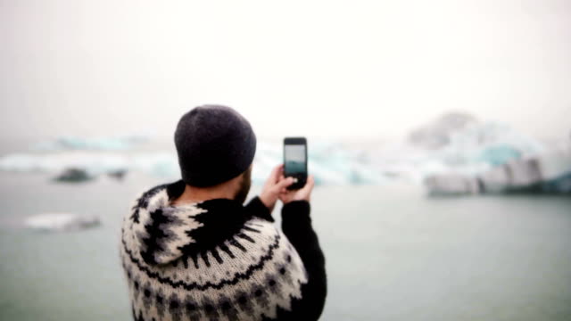 Young-stylish-man-standing-on-the-shore-in-Jokulsalon-ice-lagoon-in-Iceland-and-taking-photos-on-smartphone