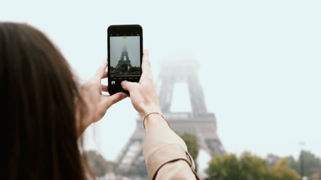 Young-beautiful-woman-walking-in-Paris,-France-and-taking-photos-of-Eiffel-tower-in-fog-on-smartphone-or-mobile-phone
