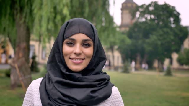 Junges-schönes-muslimische-Mädchen-Hijab-ist-stehend-und-lächelnd-in-tagsüber-bei-schlechtem-Wetter,-gerade-in-die-Kamera,-aufbauend-auf-Hintergrund,-Religiuos-Konzept,-der-Hintergrund-jedoch-unscharf