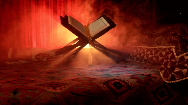 Open-holy-book-of-Muslims-on-stand-on-eastern-carpet-with-dark-toned-foggy-background.-Muslim-religion-concept.-Selective-focus.-Slider-shot.